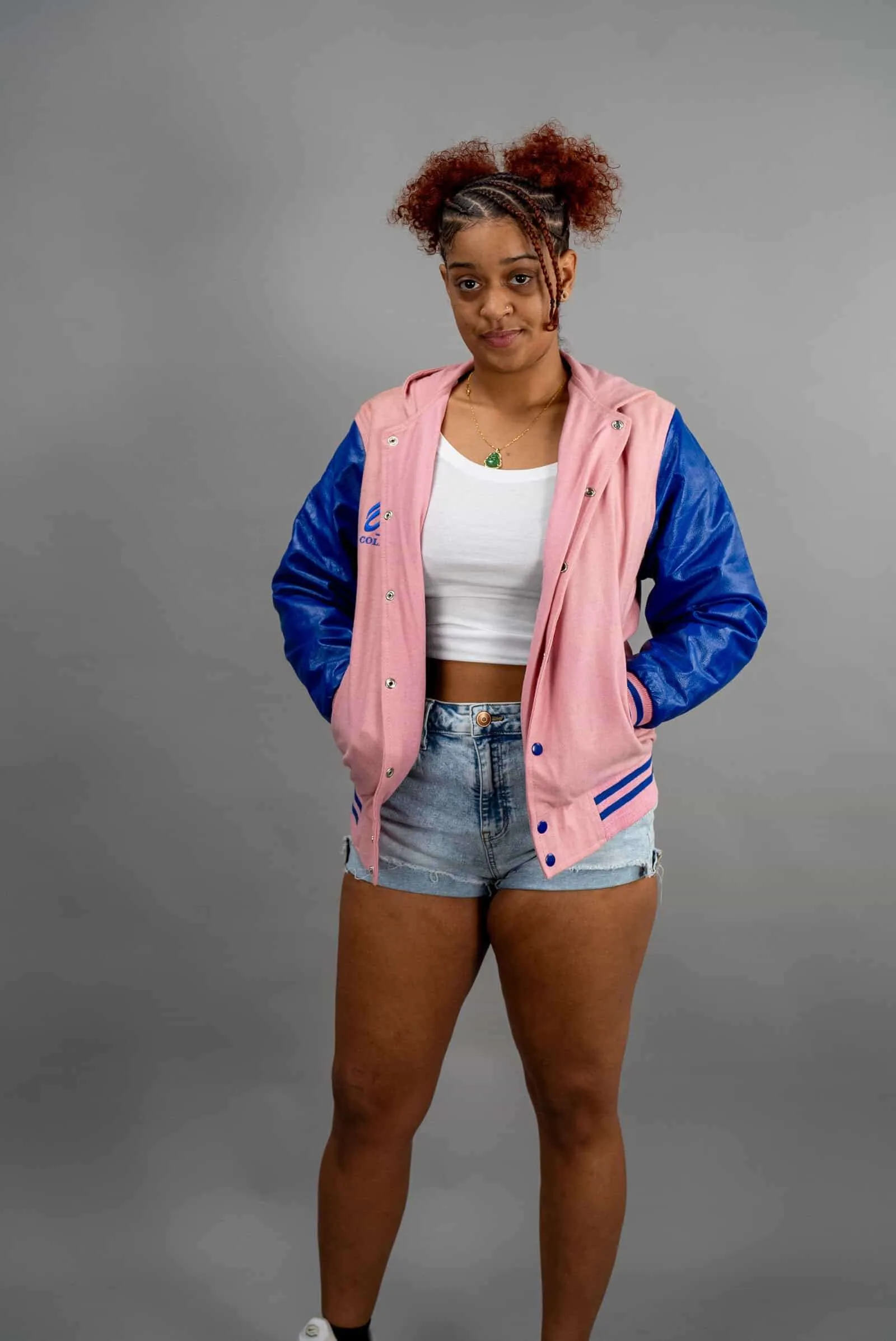 Young woman in casual pink and blue varsity jacket with denim shorts against gray background.